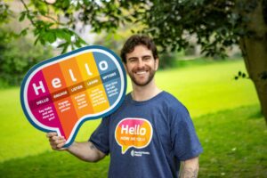 Eric Roberts stands outdoors holding a colourful Hello speech-bubble sign and wearing a Hello, How Are You? campaign T-shirt.